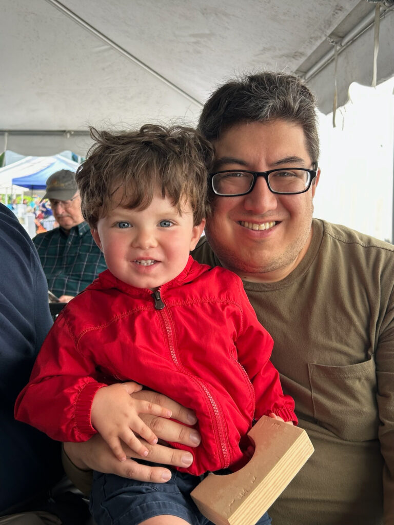 Latino man with dark hair - some grays in the front - and black-rimmed glasses sitting at an outdoor tented venue with a two year old kid with bright blue eyes and wearing a red jacket. Both are smiling - the kid is probably saying "cheese." The kid is holding a wooden block shaped like an arch.