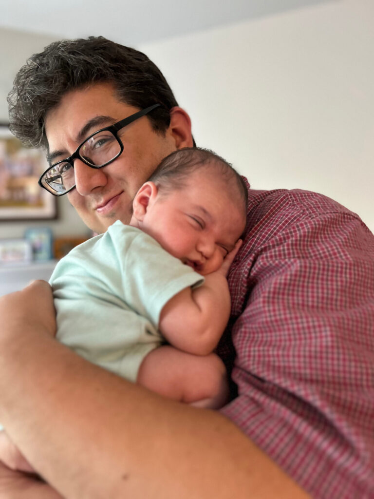 Latino man with dark hair - some grays in the front - and black-rimmed glasses holding a tiny baby in a green onesie. The baby is scrunched up with shut eyes, pressing their cheek against their tiny hand, seeking comfort in sleep. The man has the most contended and self-assured closed-lip smile we've ever seen on him.